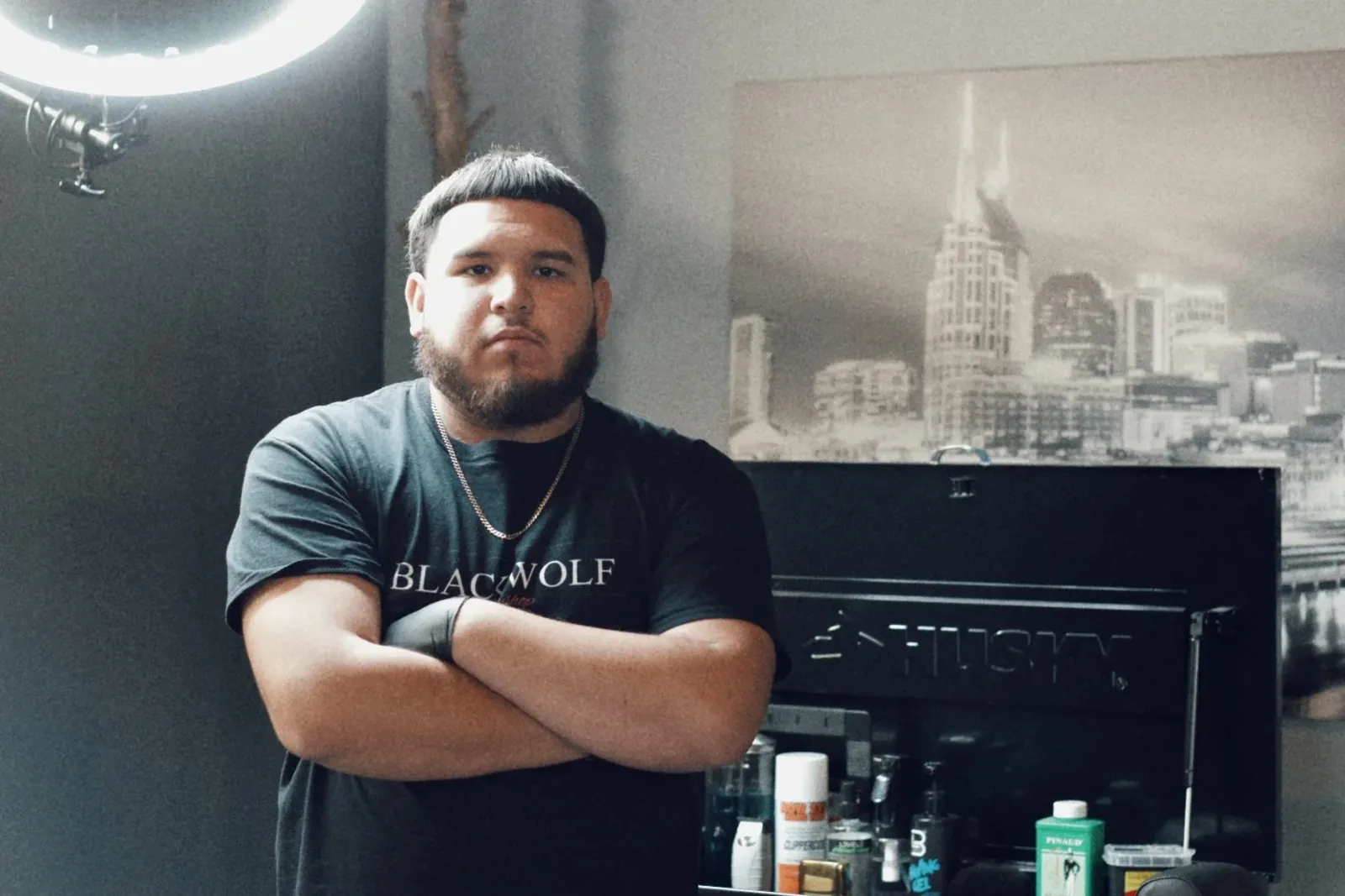 A barber in a Blackwolf T-shirt, arms crossed, beside a toolbox of products and a Nashville skyline print