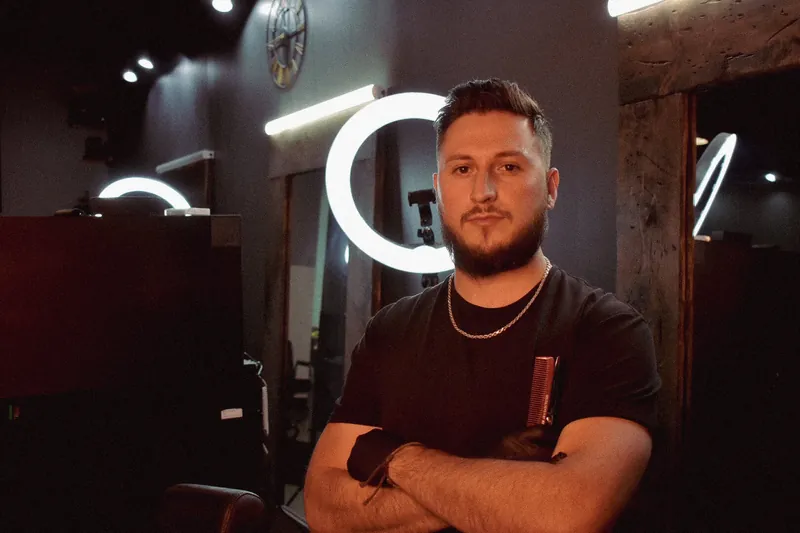 Blackwolf barber with arms crossed beside a wood-framed mirror and ring light