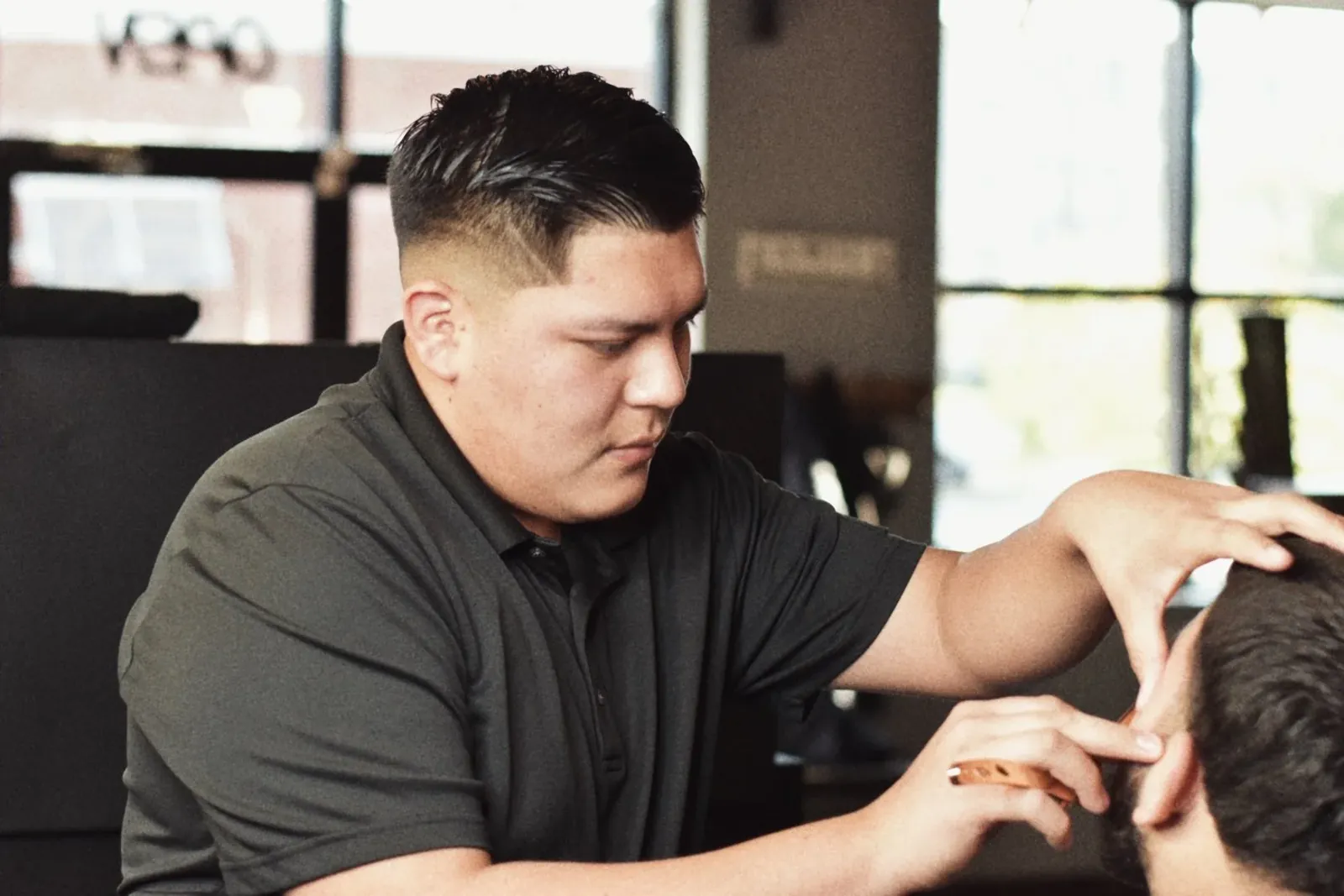 Barber in a dark polo shaping the hair above a client's ear by the shop window
