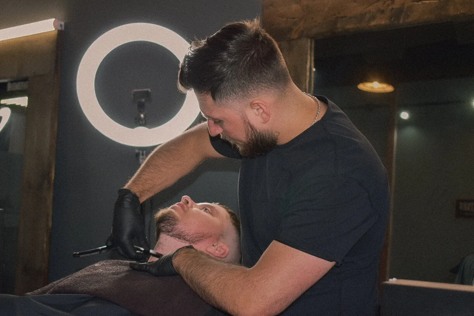Barber in black gloves lining a reclined client's beard with a razor under a ring light